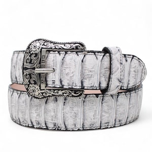 May include: A white belt with a textured pattern and a silver buckle. The buckle has an ornate design and the word "TANNER" is visible. The belt is made of a material that resembles alligator or crocodile skin.