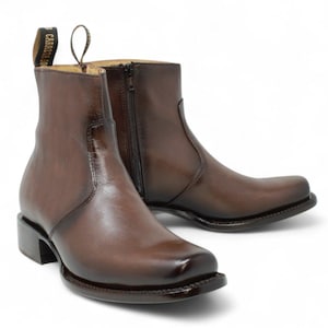 May include: Brown leather ankle boots with a square toe and a side zipper. The boots have a small heel and a pull tab at the back. The brand name "Carreta 200" is visible on the pull tab.