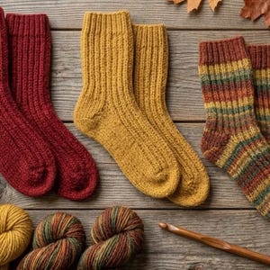 Può includere: Calzini lavorati a mano a maglia in vari colori, tra cui rosso, giallo e motivi a righe, sono disposti su una superficie di legno rustico. L'immagine include anche gomitoli di lana, un uncinetto e una tazza di tè, suggerendo un tema artigianale e accogliente.