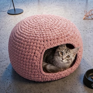 May include: A grey and white cat is nestled inside a pink, round, crocheted cat bed. A black cat food bowl with a paw print and a copper wire mouse toy are nearby. A black lamp and city skyline are in the background.