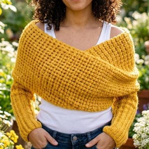 May include: A mustard yellow knitted shrug with long sleeves. The shrug is worn over a white vest top and blue jeans. The background shows a garden with green foliage and flowers.