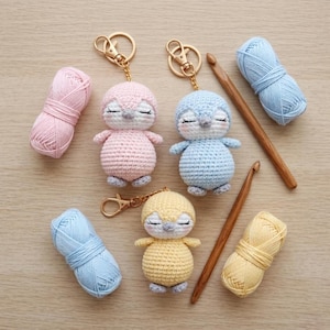 Può includere: Portachiavi a forma di pinguino all'uncinetto fatti a mano in rosa, blu e giallo, con anelli dorati. Circondati da gomitoli di filato coordinati e uncinetti in legno. I pinguini hanno gli occhi chiusi, ideali per gli artigiani.
