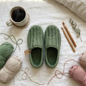 May include: Handmade green crochet slippers with a ribbed design, displayed with yarn, crochet hooks, and a cup of coffee. The slippers are a cozy, comfortable design, perfect for indoor wear. The slippers are approximately 10 inches long.