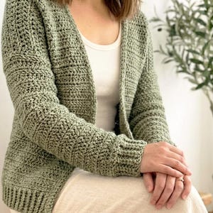 Può includere: Un cardigan all'uncinetto verde salvia con maniche lunghe. Il maglione aperto ha un motivo a punto strutturato e polsini a coste. Il cardigan è indossato sopra una canotta bianca e una gonna color crema.