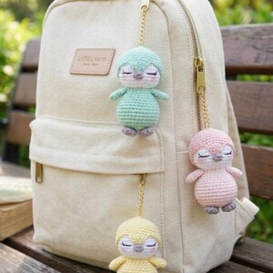 Può includere: Uno zaino beige con cerniere dorate e tre portachiavi a forma di pinguino all'uncinetto in verde menta, rosa e giallo. I pinguini hanno gli occhi chiusi e sono attaccati allo zaino con catene dorate. Lo zaino ha un'etichetta in pelle con la scritta "LITELYarn".