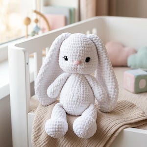 Può includere: Un coniglietto bianco all'uncinetto con lunghe orecchie flosce, occhi neri e naso rosa. Il peluche è seduto su una coperta beige all'interno di una culla bianca. L'articolo è fatto a mano.