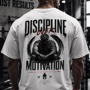 Può includere: T-shirt bianca con la scritta "DISCIPLINE OVER MOTIVATION" in un font invecchiato, con una grafica di una figura seduta. La maglietta include anche il testo "BUILT NOT GIVEN" e la frase "NO EXCUSES, NO SHORTCUTS, JUST CONSISTENCY".
