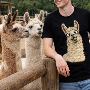 May include: Black t-shirt featuring a cartoon llama with a goofy expression. The shirt is worn by a person standing near two llamas in a field. The llamas are brown and white, and the background includes a wooden fence and trees.