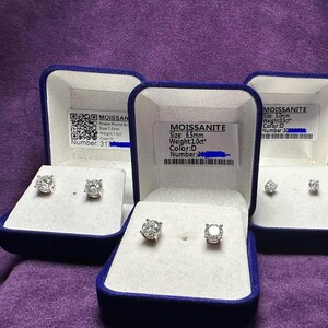 May include: Several pairs of moissanite stud earrings displayed in blue velvet boxes. Each earring is a round cut stone. The boxes have labels with details including size, weight, and color.