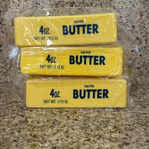 May include: Three yellow butter sticks, each wrapped in clear plastic. The packaging displays the text "SALTED BUTTER", "4oz", and "NET WT. (113 G)". The butter is stacked on a speckled countertop.