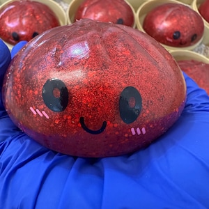Könnte beinhalten: Ein rundes, durchscheinendes rotes Quetschspielzeug, gefüllt mit feinem rotem Glitzer. Die Oberfläche zeigt ein aufgedrucktes schwarzes Gesicht mit zwei großen kreisförmigen Augen, einem geschwungenen lächelnden Mund und drei kleinen rosa Strichen.