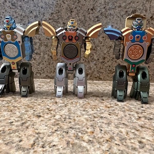 May include: Three colorful robot action figures with articulated limbs and wings. The robots have a mix of metallic colors, including blue, gold, and silver. Each robot has a circular design on its chest.