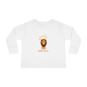 Op de afbeelding: Wit shirt met lange mouwen met een cartoon leeuw met een gouden kroon en de tekst "FUTURE LION KING" in het rood. De leeuw heeft een bruine manen en lacht. De kroon heeft kleurrijke accenten.