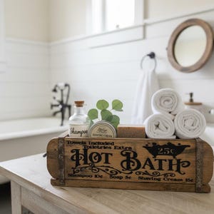 May include: A rectangular wooden crate with metal accents featuring the black printed text Hot Bath, Towels Included, Toiletries Extra, and price listings. It holds a glass shampoo bottle, a shaving cream tin, a bar of soap, and three white towels.