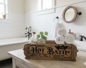 May include: A rectangular wooden crate with metal accents featuring the black printed text Hot Bath, Towels Included, Toiletries Extra, and price listings. It holds a glass shampoo bottle, a shaving cream tin, a bar of soap, and three white towels.