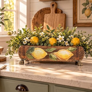 May include: A rectangular wooden planter box with a rustic brown finish, featuring a painted design of two yellow lemons with green leaves on the front. The planter stands on four small, ornate metal feet and is filled with green foliage and white flowers.