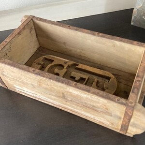 May include: A rectangular wooden brick mold featuring a weathered light brown finish with rusted metal bands securing the sides. The interior base contains a carved wooden insert with the letters G and F clearly visible in a raised relief design.