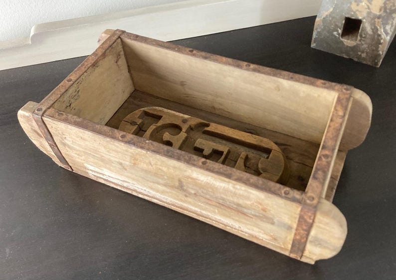 May include: A rectangular wooden brick mold featuring a weathered light brown finish with rusted metal bands securing the sides. The interior base contains a carved wooden insert with the letters G and F clearly visible in a raised relief design.