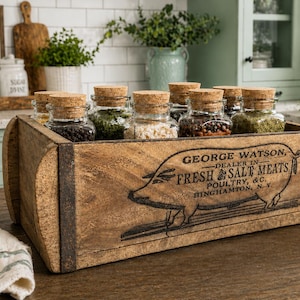 May include: A rectangular wooden crate with dark metal corner accents, featuring a black printed graphic of a pig and the text GEORGE WATSON, DEALER IN FRESH SALT MEATS, POULTRY, & C., BINGHAMTON, N.Y. The crate contains several glass spice jars.