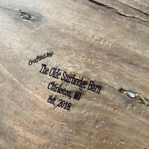 May include: A light brown wooden surface with a natural grain and a small knot. The text is printed in black ink and reads: Crafted by: The Olde Sturbridge Barn, Clarkston, MI, est. 2018. The wood features a visible texture and a small splintered area.