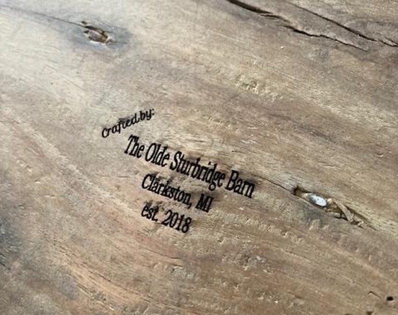 May include: A light brown wooden surface with a natural grain and a small knot. The text is printed in black ink and reads: Crafted by: The Olde Sturbridge Barn, Clarkston, MI, est. 2018. The wood features a visible texture and a small splintered area.