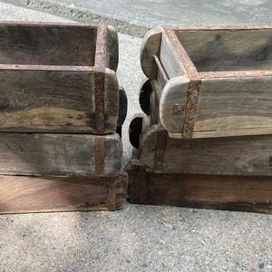 May include: A set of four rectangular wooden brick molds stacked in two columns. Each mold features weathered, dark brown wood grain and is reinforced with rusted metal bands around the edges, showing a hollow interior and open top design.