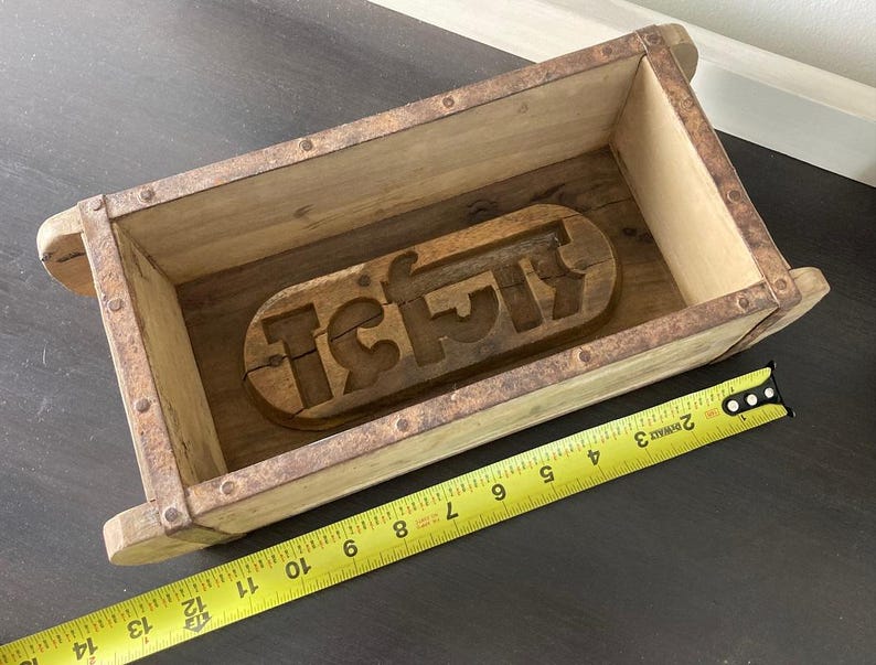 May include: A rectangular wooden mold with rusted metal edges and a carved wooden insert featuring Hindi script at the base. A yellow measuring tape is positioned along the front, showing the mold length is approximately 13.5 inches long.