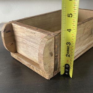 May include: A rectangular wooden brick mold with a light brown natural finish and a weathered texture. A yellow DeWalt measuring tape is positioned vertically against the side, showing a height measurement of approximately 5.5 inches.
