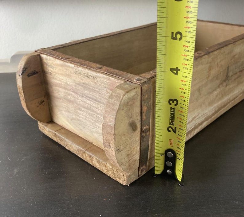 May include: A rectangular wooden brick mold with a light brown natural finish and a weathered texture. A yellow DeWalt measuring tape is positioned vertically against the side, showing a height measurement of approximately 5.5 inches.