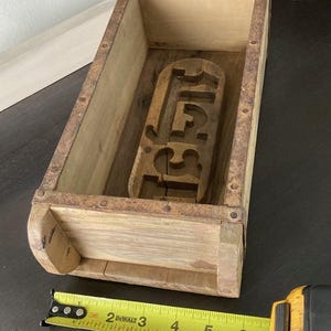 May include: A rectangular wooden brick mold with rusted metal edges and a carved interior design. A yellow DeWalt measuring tape is positioned along the base, showing the mold length is approximately 6.5 inches long and 3 inches wide.