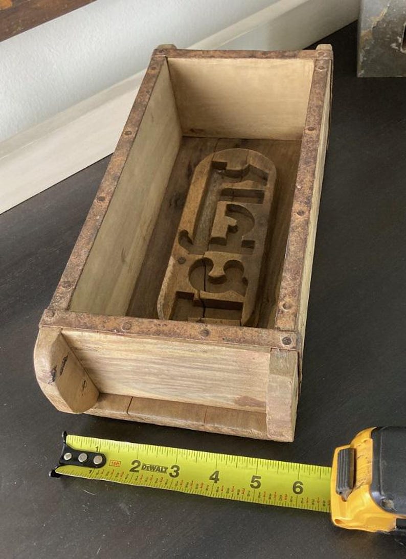 May include: A rectangular wooden brick mold with rusted metal edges and a carved interior design. A yellow DeWalt measuring tape is positioned along the base, showing the mold length is approximately 6.5 inches long and 3 inches wide.