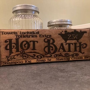 May include: A rectangular wooden brick mold with metal bands, featuring the burned-in text "Hot Bath," "Towels Included," "Toiletries Extra," and prices for shampoo, soap, and shaving cream. Two clear glass jars with metal lids sit behind the mold.