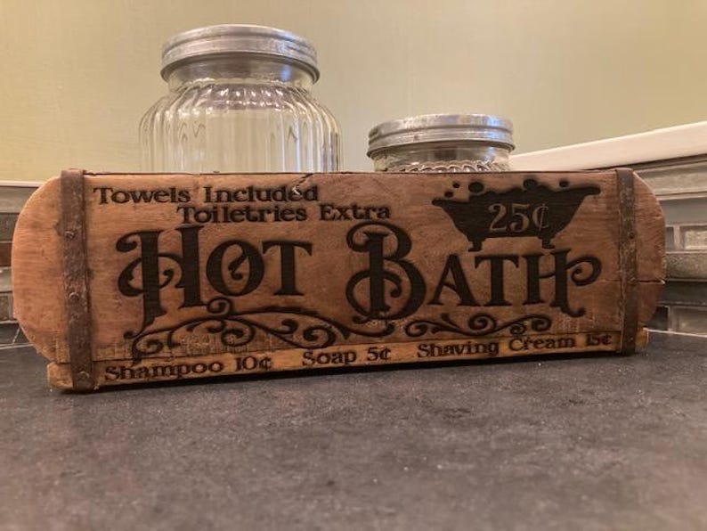 May include: A rectangular wooden brick mold with metal bands, featuring the burned-in text "Hot Bath," "Towels Included," "Toiletries Extra," and prices for shampoo, soap, and shaving cream. Two clear glass jars with metal lids sit behind the mold.