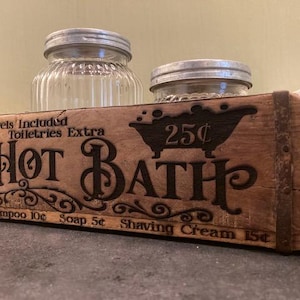 May include: A rectangular wooden box with metal bands featuring the text "Hot Bath" and a bathtub graphic. The text includes "Towels Included," "Toiletries Extra," "25&cent;," "Shampoo 10&cent;," "Soap 5&cent;," and "Shaving Cream 15&cent;." Two glass jars sit behind it.