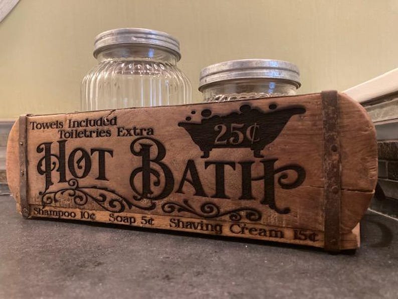 May include: A rectangular wooden box with metal bands featuring the text "Hot Bath" and a bathtub graphic. The text includes "Towels Included," "Toiletries Extra," "25&cent;," "Shampoo 10&cent;," "Soap 5&cent;," and "Shaving Cream 15&cent;." Two glass jars sit behind it.