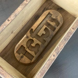 May include: A rectangular wooden box containing a carved wooden mold with the letters ISH. The mold is light brown with a visible wood grain and a vertical crack running through the letter I. The box has light-colored wooden sides and a dark base.