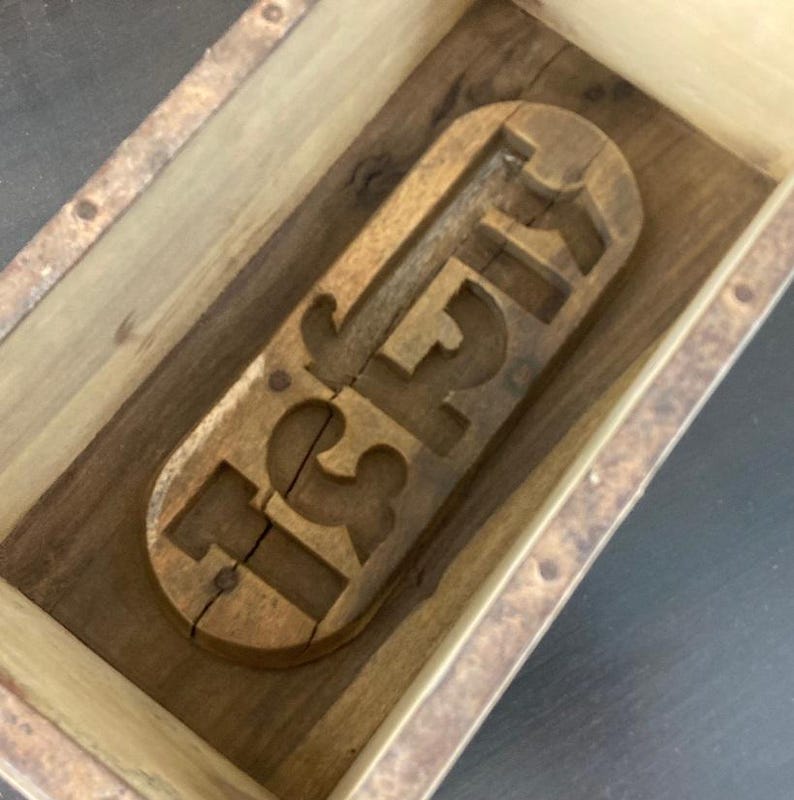 May include: A rectangular wooden box containing a carved wooden mold with the letters ISH. The mold is light brown with a visible wood grain and a vertical crack running through the letter I. The box has light-colored wooden sides and a dark base.