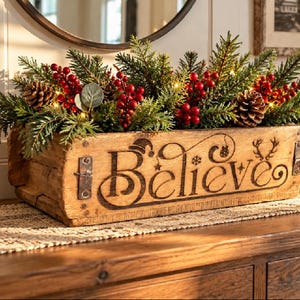May include: A rectangular wooden box with metal accents featuring the word Believe burned into the front. The box is filled with green pine branches, red berries, pinecones, and small warm white string lights for a holiday display.