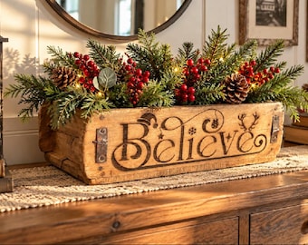 May include: A rectangular wooden box with metal accents featuring the word Believe burned into the front. The box is filled with green pine branches, red berries, pinecones, and small warm white string lights for a holiday display.