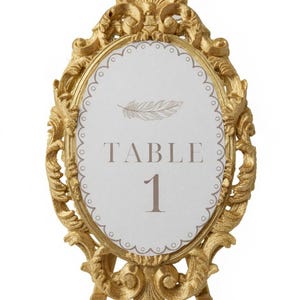 Könnte beinhalten: Ein ovales Tischnummernschild mit einem verzierten Goldrahmen mit komplexen Schnörkeln und einem weißen Einsatz. Der Einsatz zeigt eine hellbraune Federillustration über dem Text "TABLE 1" in einer einfachen, dunklen Serifenschrift.