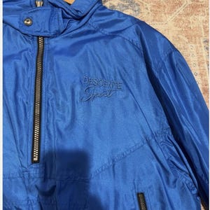 May include: Bright blue athletic jacket with a high collar and a black zipper. The jacket has the text "DESCENTE Sport" embroidered on the chest. The jacket is hanging on a wooden hanger.
