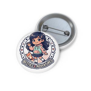 May include: White button badge featuring a cartoon girl waving, a fox, and the text "Besties & Adventures." The girl is wearing a pink hoodie and blue skirt. The badge has a metal pin on the back.