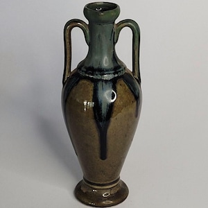 May include: A ceramic amphora vase featuring a narrow neck and two symmetrical handles. The body displays a mottled brown base glaze with a dark blue-green drip pattern cascading from the rim down the neck and onto the upper shoulder area.