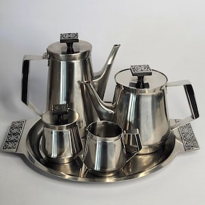 May include: A stainless steel tea and coffee service set consisting of a tall coffee pot, a shorter teapot, a creamer, and a sugar bowl with a lid, all arranged on a matching round serving tray with decorative black and white floral side panels.
