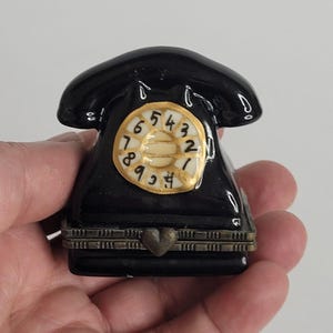 May include: A small black ceramic trinket box shaped like a vintage rotary telephone. The piece features a gold-rimmed circular dial with black numbers and a metallic hinge closure with a small heart-shaped clasp at the front centre.