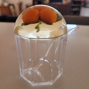 May include: A clear, faceted acrylic sugar bowl with a matching lid and spoon. The lid features an embedded decorative element consisting of three small, round orange fruits and green leaves encased within the transparent, dome-shaped top.