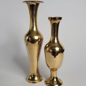 May include: Two polished brass vases with a reflective gold-toned finish. The taller vase features a slender, elongated neck and a flared rim, while the shorter vase has a wider, bulbous body and a flared base, both standing on circular pedestals.