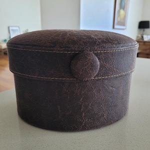 May include: A circular, brown leather box with a button fastening. The box has a textured finish and a stitched seam around the top and base. The box is placed on a light-coloured surface.