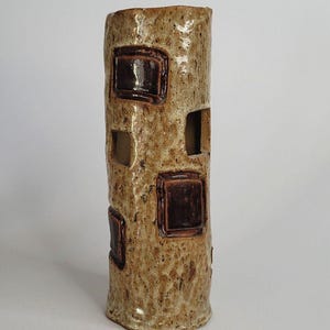May include: A cylindrical ceramic vase with a textured, speckled tan surface. The exterior features several raised, dark brown square accents and open rectangular cutouts distributed across the body, creating a hollow, architectural appearance.