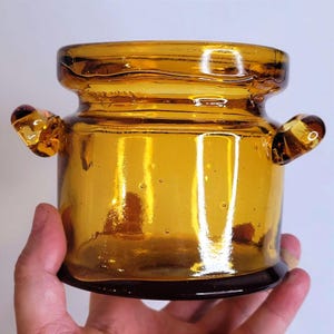 May include: A small, amber-coloured glass jar with a wide mouth and two small, rounded handles on opposite sides. The glass features a smooth, translucent finish with visible air bubbles throughout the body and a slightly darker, thicker base.